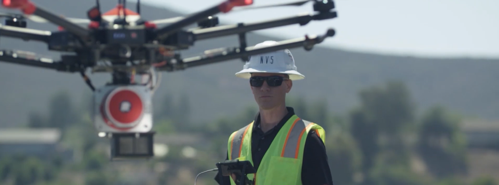 Man wearing white NV5 hard hat with safety vest piloting drone for aerial drone survey